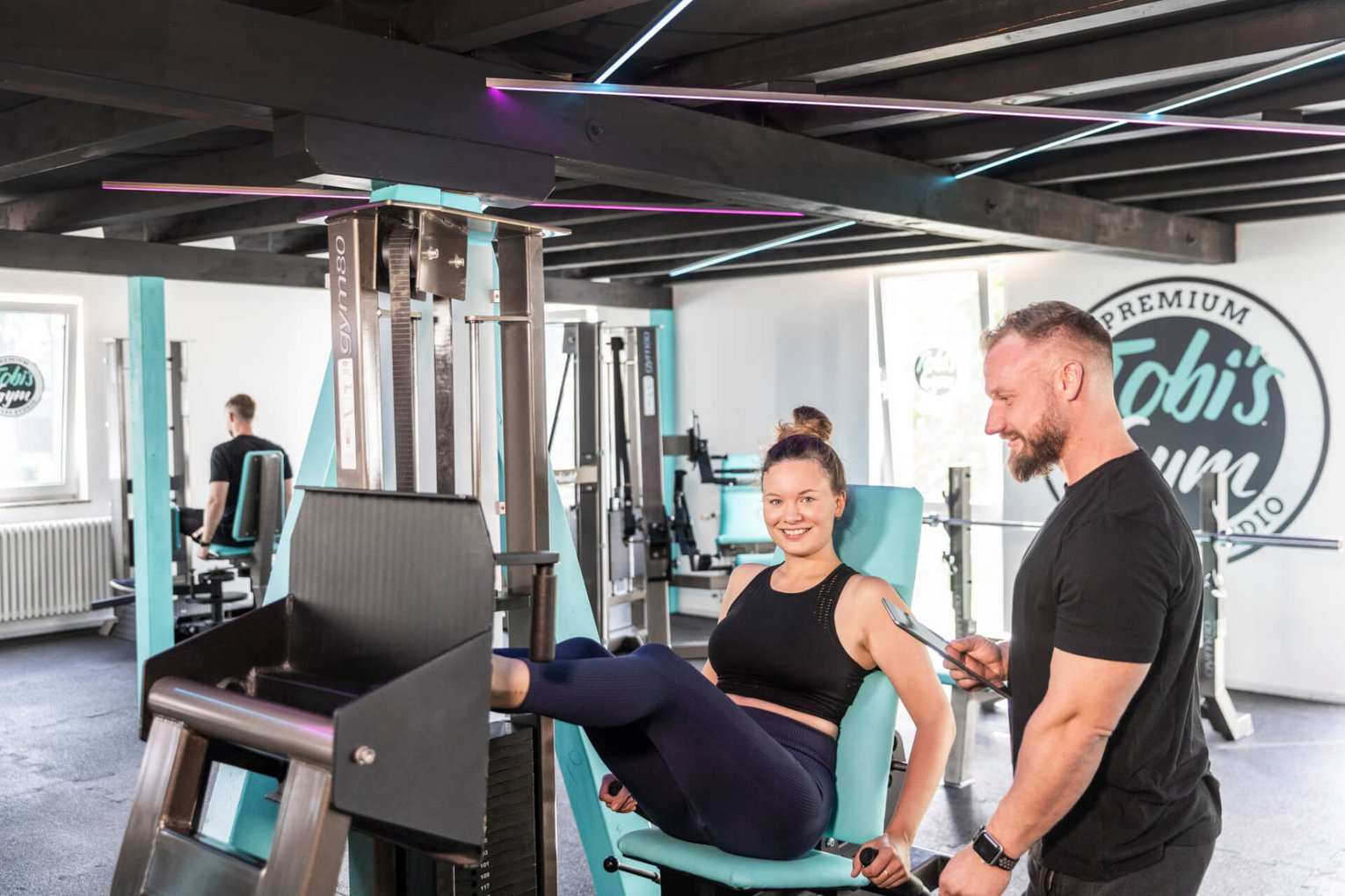 Tobi's Gym Beinpresse Sportlerin trainiert auf der Beinpresse in Tobi's Gym, Trainer steht daneben